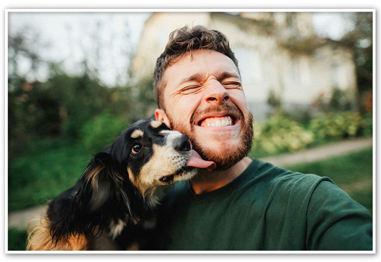 Man smiling with eyes closed as a dog licks his face outdoors.