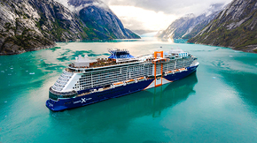 A large cruise ship sails through a turquoise fjord, flanked by steep, snowy mountains.